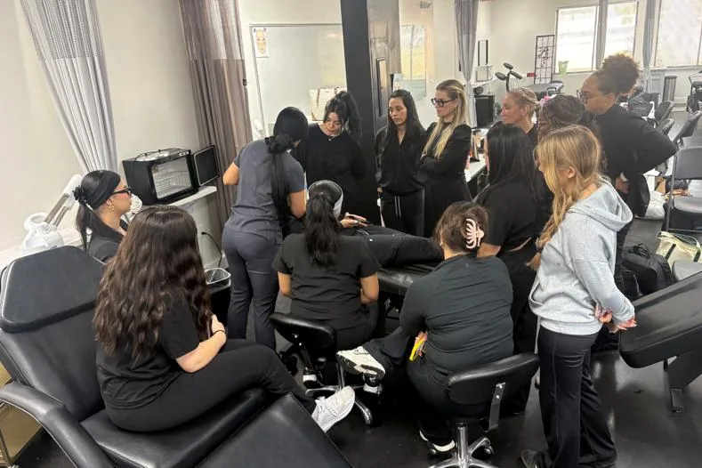 Esthetics students gathered around an instructor's treatment demo at Hyphen Beauty + Massage School in Winter Park, FL