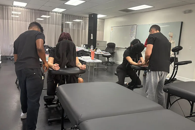 Massage students doing hands-on practice in a bright classroom at Hyphen Beauty + Massage School in Winter Park, FL