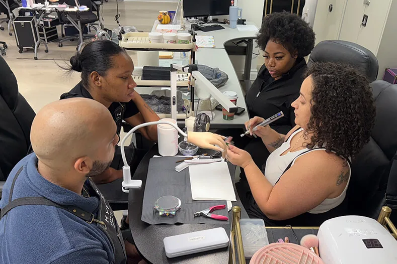 Nail tech students working on practice hands at a shared table at Hyphen Beauty + Massage School in Winter Park, FL